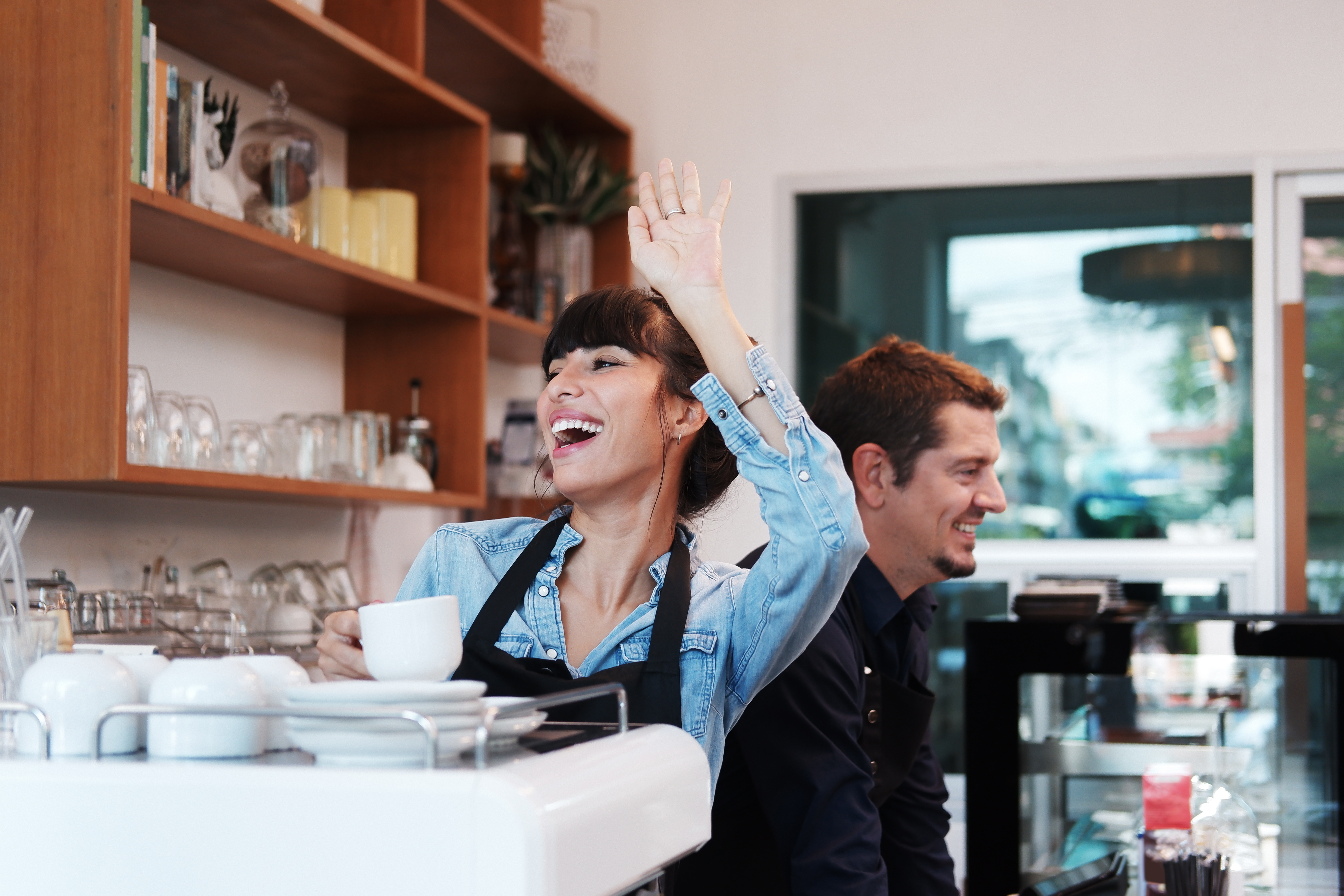 A female cafe barista smiles and waves at a person out of frame. Her male coworker smiles behind her, looking away.