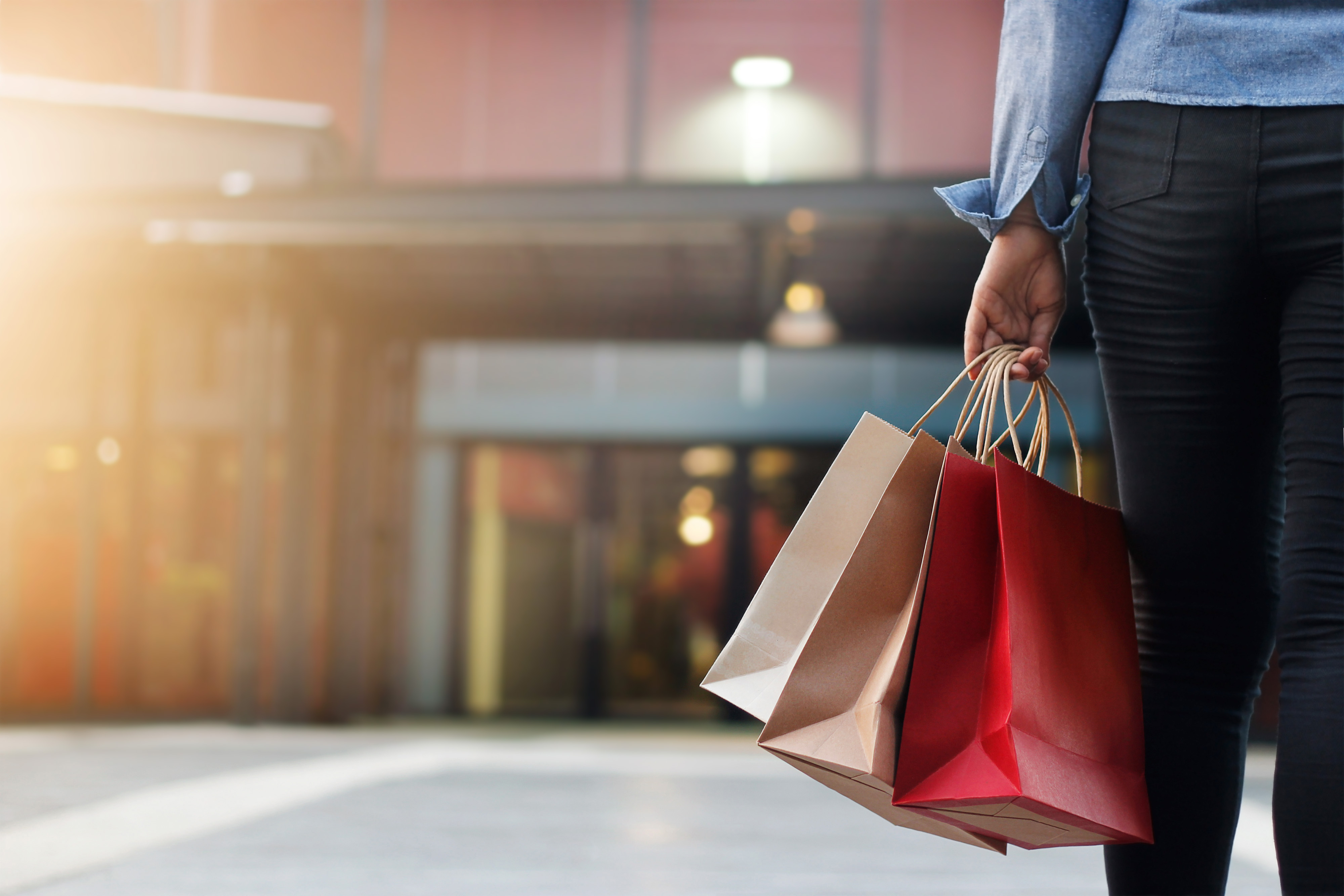 A person wearing black jeans and a blue shirt holds three paper shopping bags as they stand in front of a store.