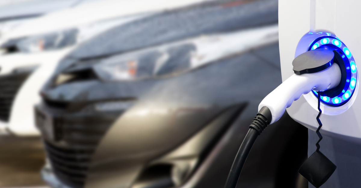 An electric vehicle charging station with the charger plugged into a blue ring and several cars parked in the background.