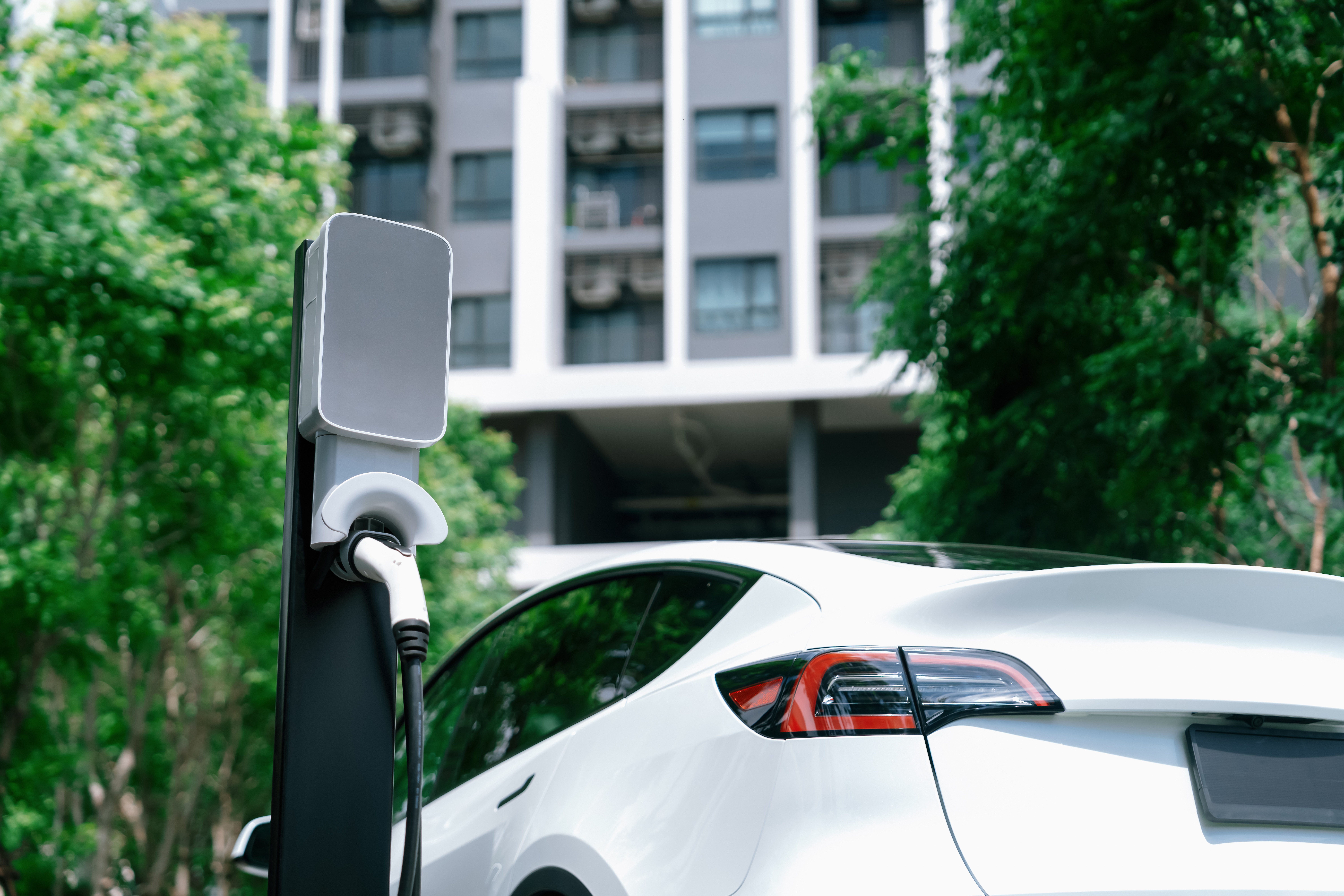 A close-up of a white electric car plugged into an EV charger in front of an apartment building with balconies.