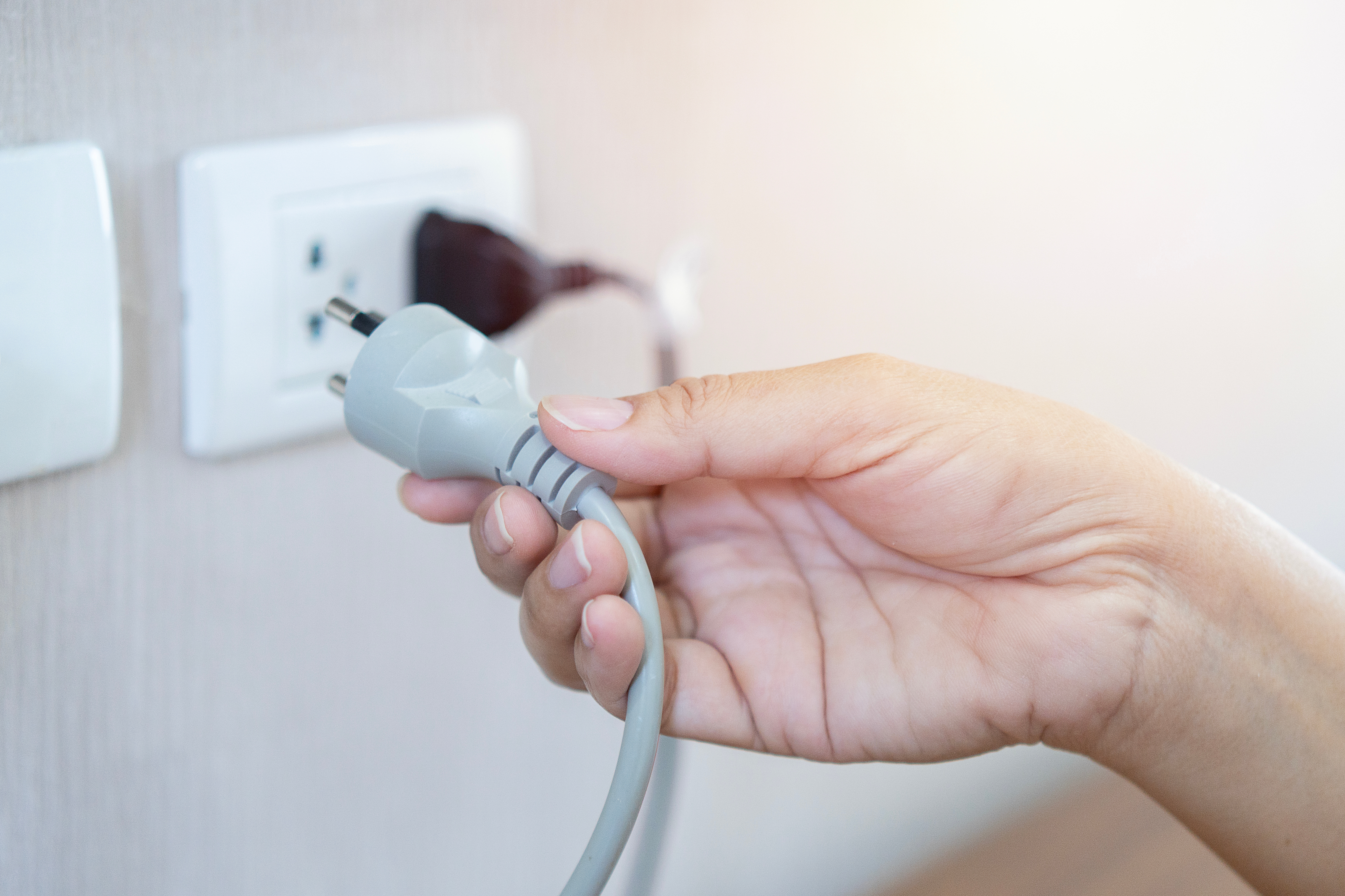 A close-up of a pale hand plugging or removing a plug on a gray extension cord from an outlet in a wall.