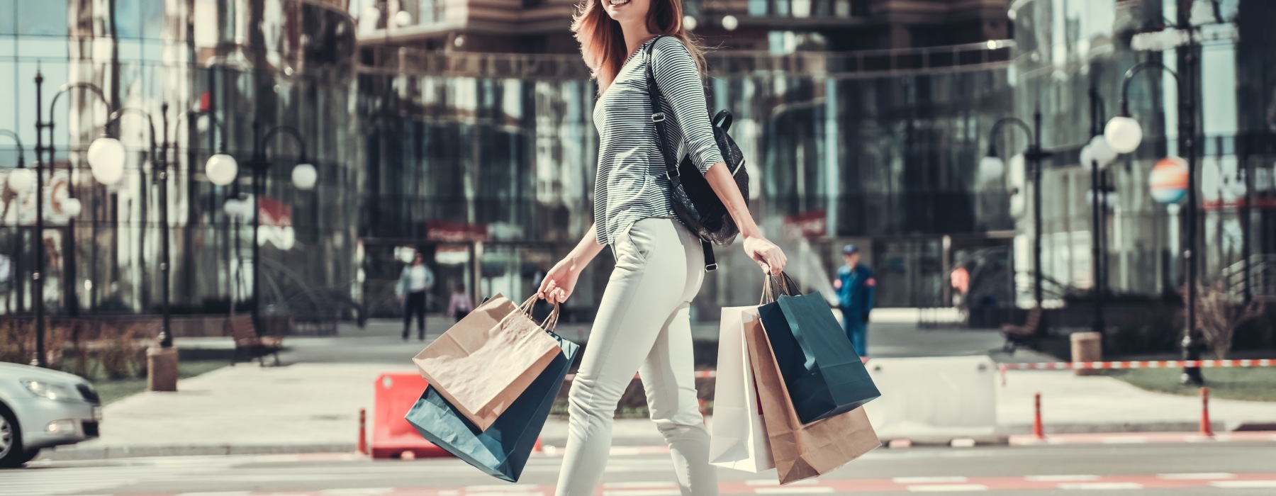 A woman carrying several colorful paper shopping bags in each hand walks across a crosswalk in the middle of an urban area.