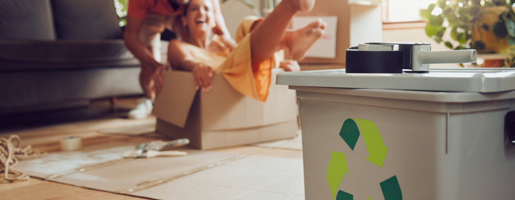 A couple moving into a new place playing in boxes in the background, with focus on a gray recycling bin in the foreground.