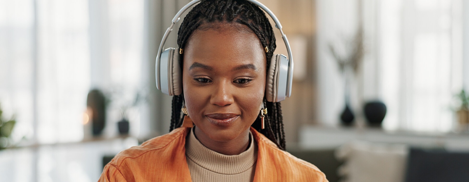 A young woman sitting in a living space with gray headphones on. She looks down at the laptop in front of her.
