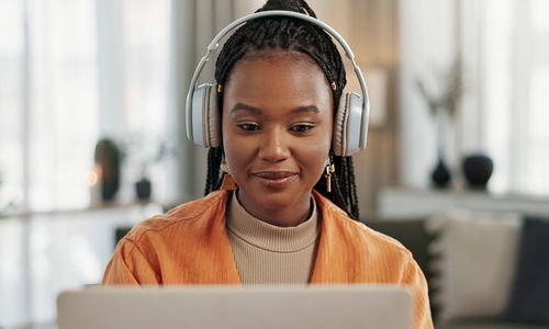 digtigardqozbllc-391030-woman-on-computer-image-blogbanner1 A young woman sitting in a living space with gray headphones on. She looks down at the laptop in front of her.