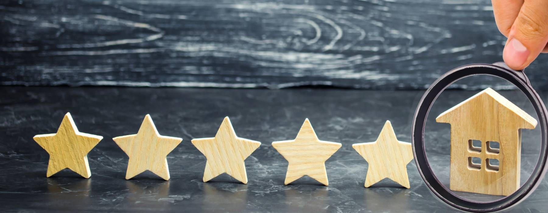 A hand holds a magnifying glass up to a wooden cutout of a house. Five wooden star cutouts line up next to it.