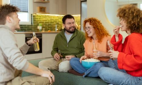 A group of friends sit on a couch in an apartment space, holding bottled drinks and sharing a bowl of popcorn.
