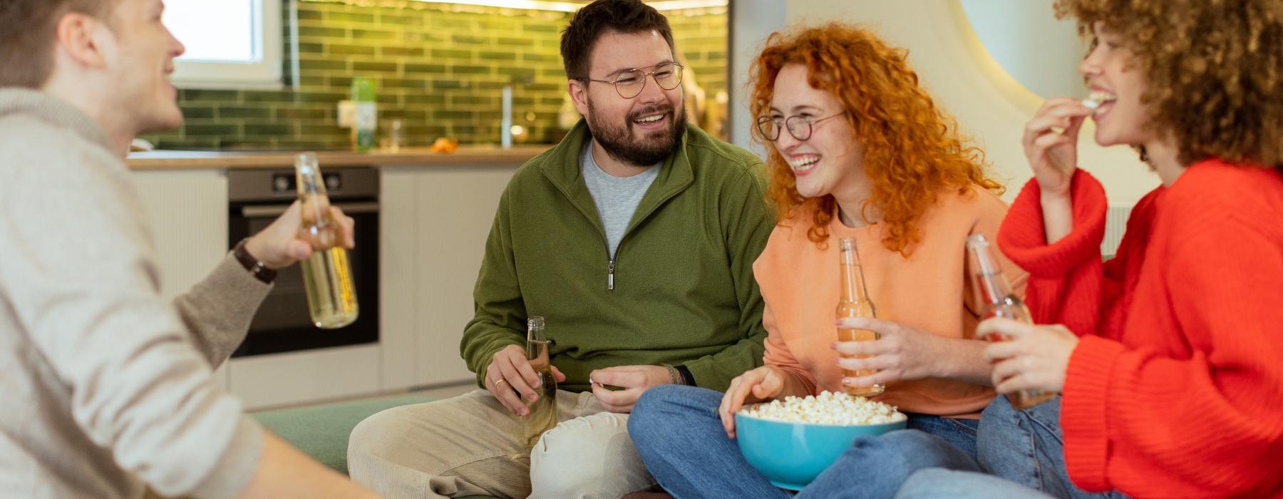 A group of friends sit on a couch in an apartment space, holding bottled drinks and sharing a bowl of popcorn.