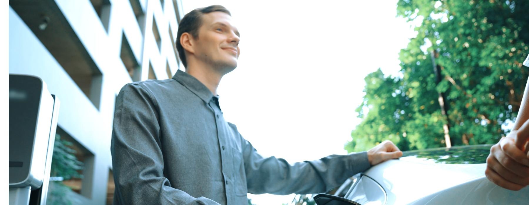 A man in a gray button-down stands in front of his electric vehicle, holding the EV charger and looking at a person out of frame.