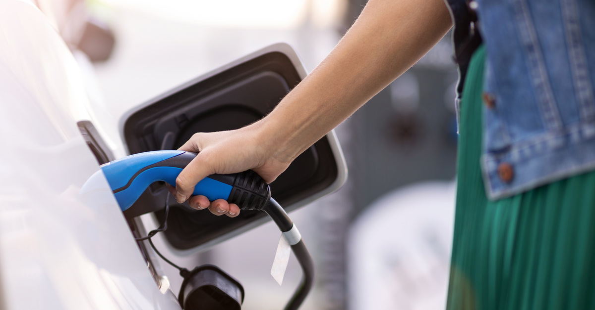 A close-up of a hand of a person wearing green and blue holding a blue connector to charge a white electric vehicle.