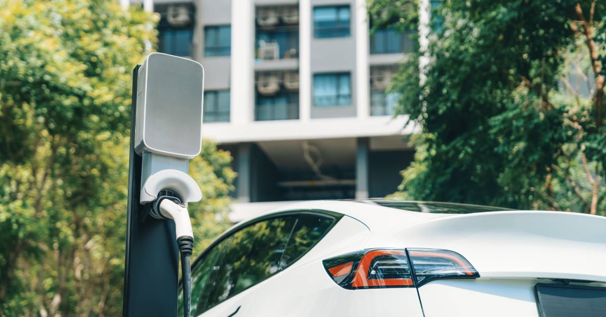 digtigardqozbllc-391034-ev-car-charging-image-a1 An EV charging cable is plugged into the station next to a white car parked outside an apartment complex.