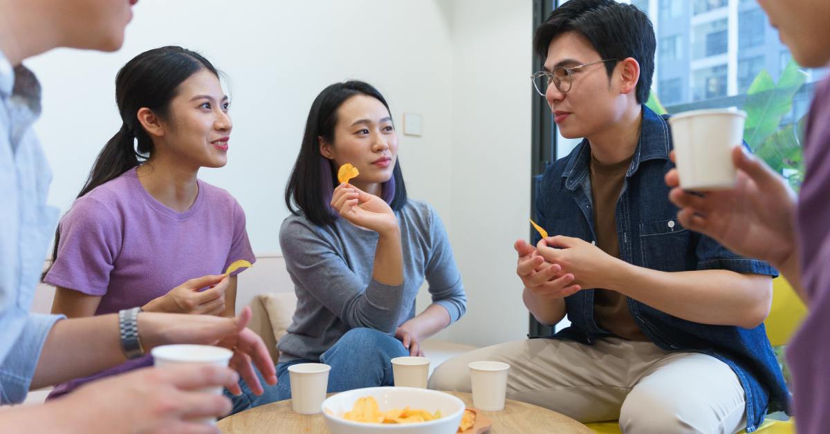 digtigardqozbllc-391038-group-of-friends-image-b1 A group of friends in mid-conversation sitting around a small table next to a window and sharing a bowl of chips.