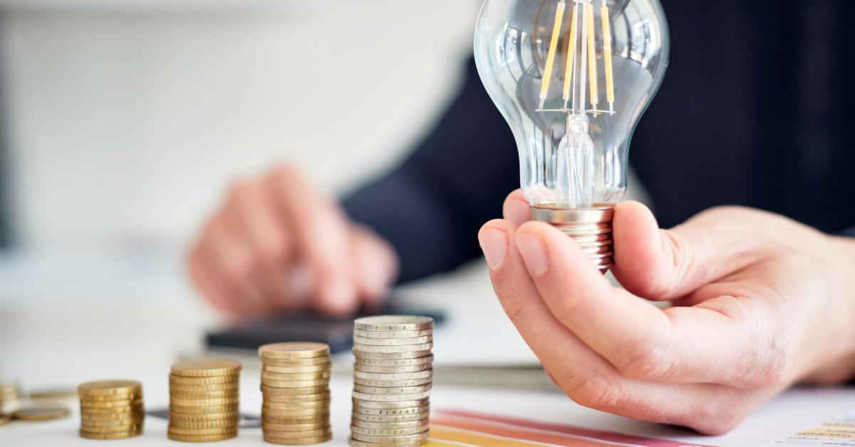 digtigardqozbllc-391044-holding-light-bulb-image-a1 A close-up of a person sitting at a desk and holding a light bulb next to ascending stacks of coins and a graph.