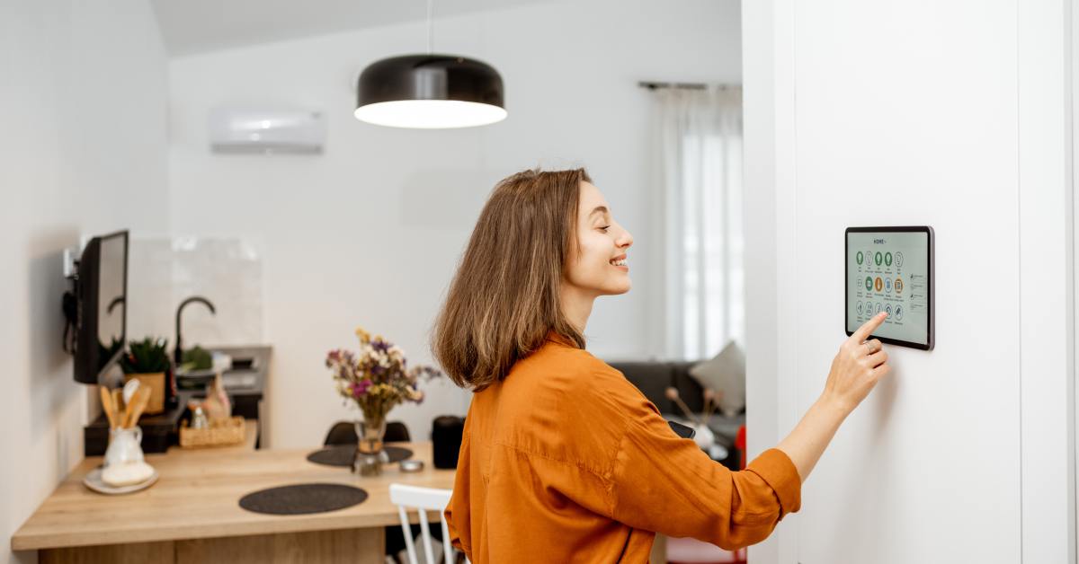 digtigardqozbllc-391044-woman-tapping-screen-image-b1 A woman wearing an orange shirt smiles as she taps the screen of a smart home panel on the white wall of a room.