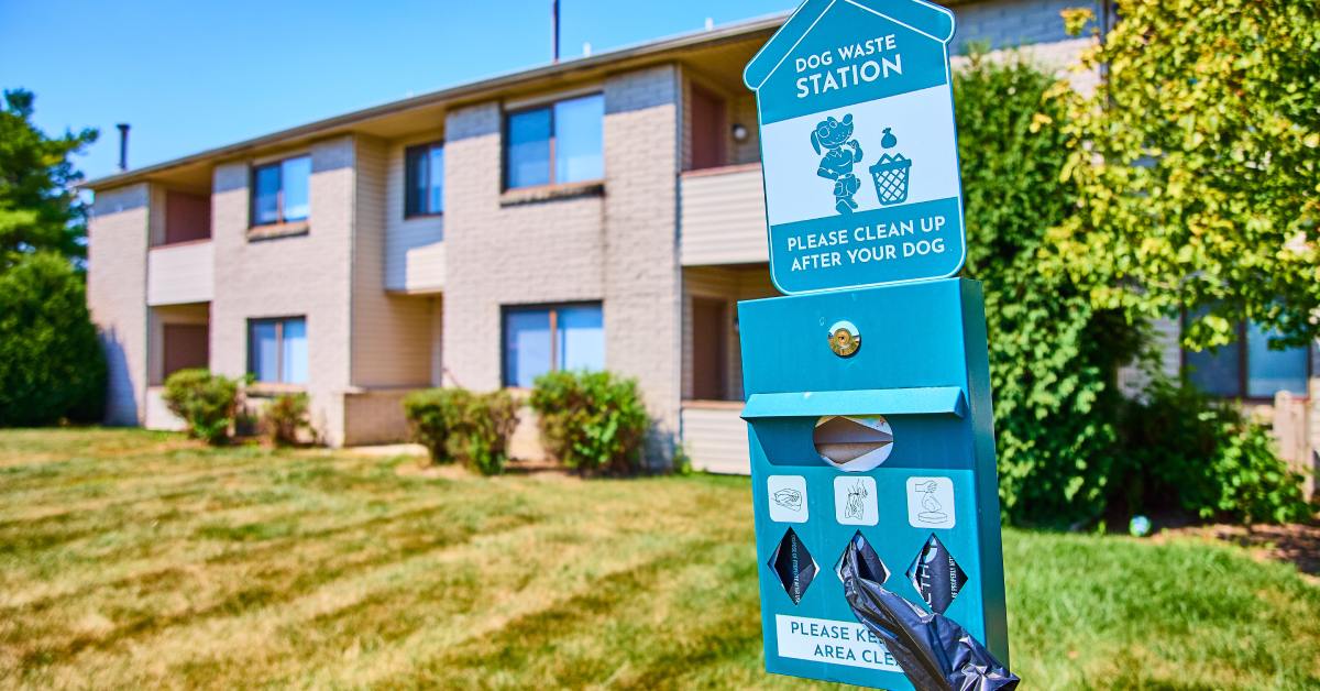 digtigardqozbllc-391046-dog-waste-station-image-b1 A close-up of a dog waste station in focus outside of an apartment complex, which is across the lawn in the background.